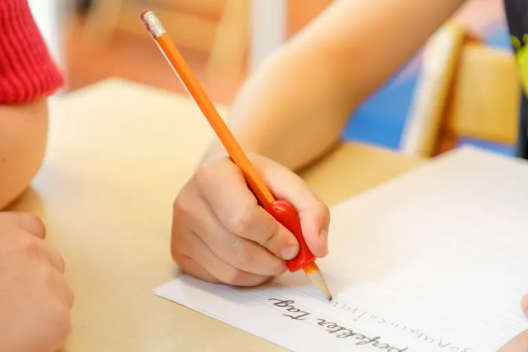 Kinderhand schreibt mit einem Stift und einer Griffhilfe auf Linienpapier