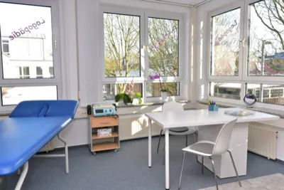 Logopädieraum mit Arbeitstisch und Therapieliege vor einer breiten Fensterfront